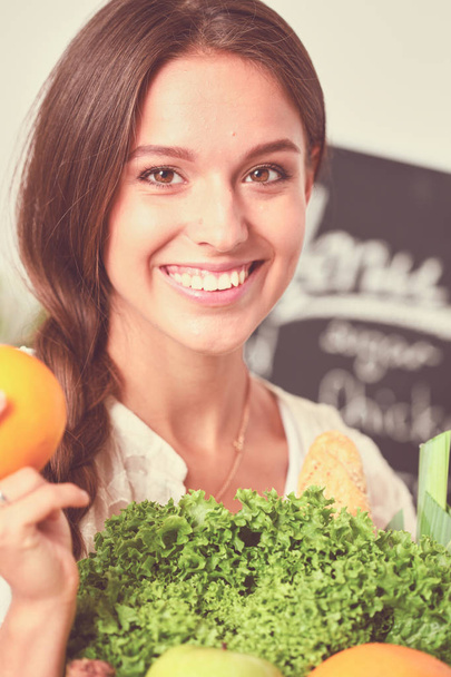 Sorrindo jovem segurando legumes de pé na cozinha. Sorrindo jovem mulher - Foto, Imagem