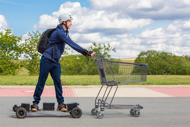 Dutch teenage boy  rides on electric skateboard pushing shopping cart outdoors - Fotó, kép