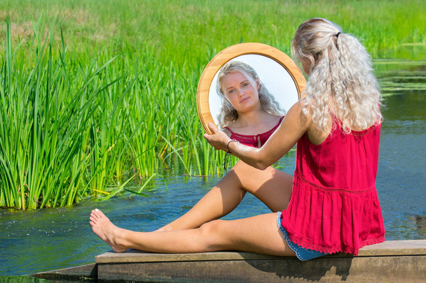 Молодая блондинка европейская женщина сидит с зеркалом у воды в природе
 - Фото, изображение