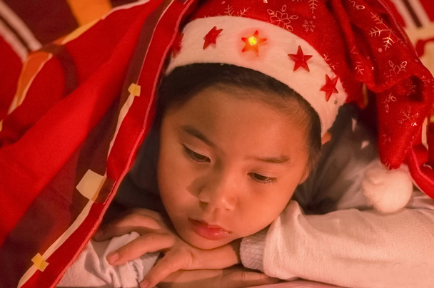 Niño llevaba un sombrero de Navidad debajo de una manta, tristemente y solo en el dormitorio en una noche fría de Navidad
. - Foto, Imagen