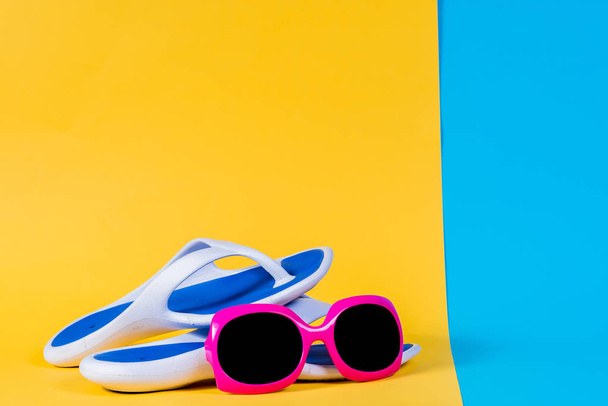 Μπλε σαγιονάρες, σανδάλια, γυαλιά ηλίου και θαλασσινών κοχυλιών, καλοκαιρινή σειρά. Το καλοκαίρι είναι προσεχείς έννοια. Minimal άποψη χώρου, μινιμαλιστικής φωτογράφησης. Κίτρινο, ροζ και μπλε παστέλ χρώματα φόντου. - Φωτογραφία, εικόνα