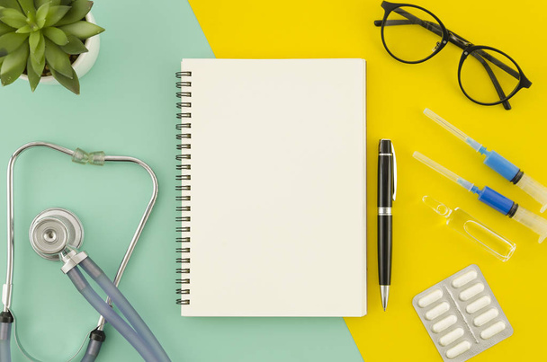 Medical or healthcare concept. Spiral blank notepad mock up on blue and yellow dual color background. Medical equipment, stethoscope. Flat lay view. - Foto, afbeelding
