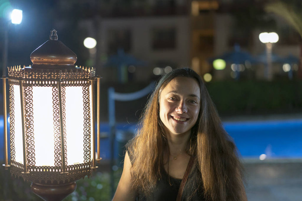 Mujer joven cerca de una gran linterna en el fondo de un hotel nocturno
. - Foto, imagen