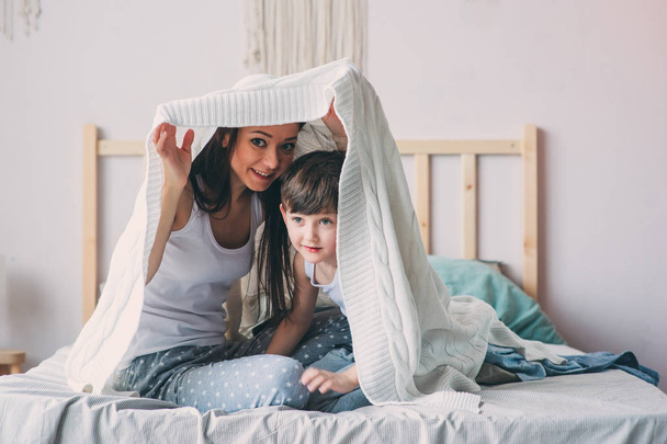 happy mother and son playing and hiding under blanket in bed in cozy weekend morning. Casual lifestyle with real family - Foto, Bild