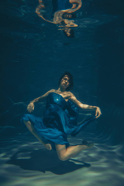 cute sporty female swims underwater as a free diver in a blue evening dress alone - Photo, image