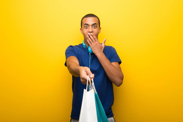 Afro-amerikai ember meglepett egy csomó bevásárlótáskák lenyomva sárga alapon kék pólóban - Fotó, kép