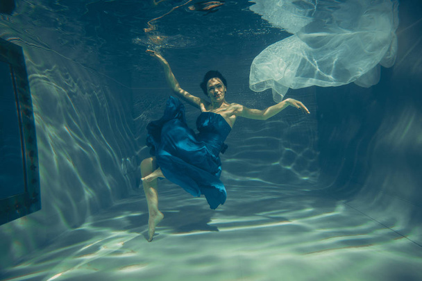 cute sporty female swims underwater as a free diver in a blue evening dress alone - Photo, Image