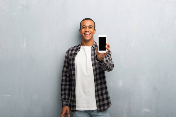junger afrikanisch-amerikanischer Mann mit kariertem Hemd blickt in die Kamera und lächelt, während er das Handy benutzt - Foto, Bild