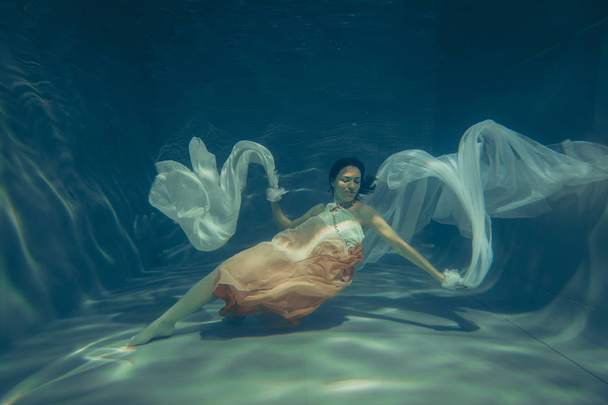 κομψό λεπτό κορίτσι υποβρύχια κολυμπά σαν έναν ελεύθερο δύτη σε ένα λευκό φόρεμα βράδυ με όμορφο ύφασμα - Φωτογραφία, εικόνα
