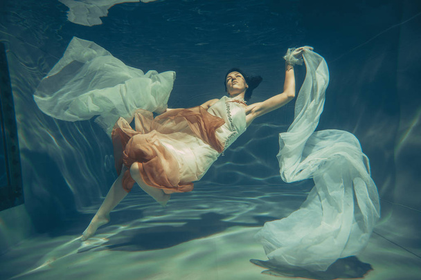 zarif ince kız su altında güzel kumaş ile beyaz gece elbisesi içinde ücretsiz bir dalgıç gibi yüzüyor - Fotoğraf, Görsel