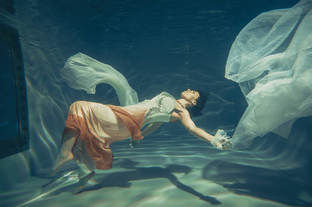 элегантная стройная девушка плавает под водой, как свободный дайвер в белом вечернем платье с красивой тканью
 - Фото, изображение