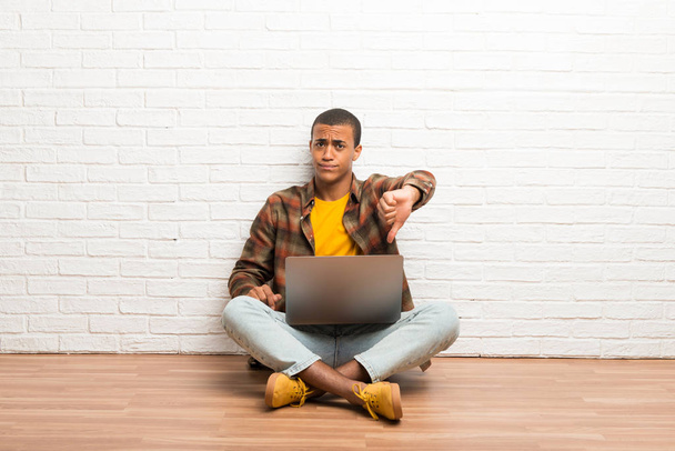 Hombre afroamericano sentado en el suelo con su portátil mostrando el pulgar hacia abajo signo con expresión negativa
 - Foto, Imagen