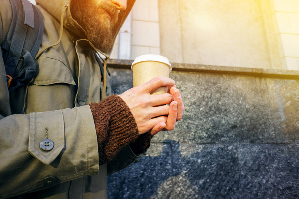 Paper cup of hot coffee in the hand of a bearded man in a jacket and a brown sweater. Warming drink in cold winter weather. Take away coffee concept. Coffee cup layout - Foto, Bild