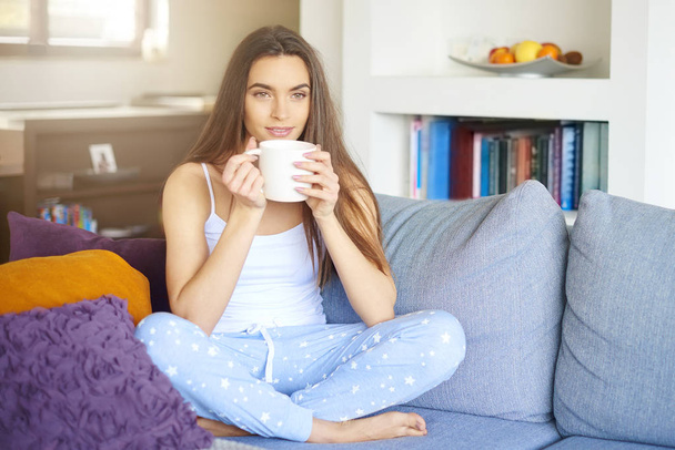 Shot of young pretty woman wearing pajamas and drinking tea while relaxing on sofa early morning. - Photo, image