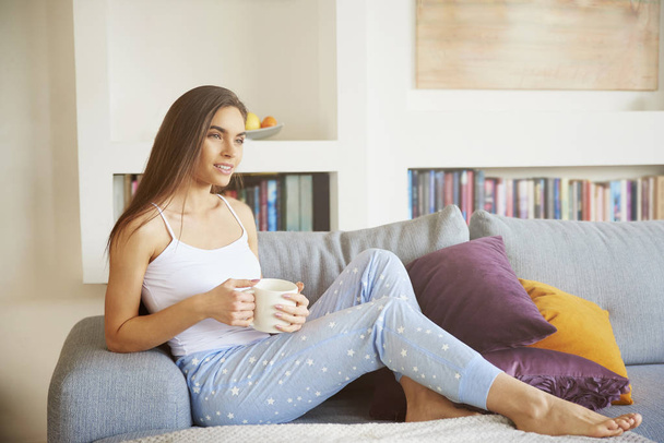 Shot of young pretty woman wearing pajamas and drinking tea while relaxing on sofa early morning. - Fotó, kép