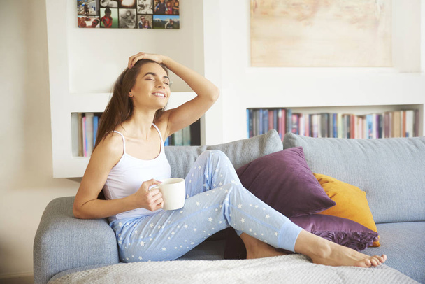 Full length shot of attractive young woman wearing pajamas and drinking tea while relaxing on the sofa in her apartment.  - Foto, Imagem