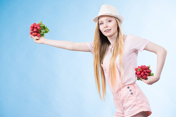 Positive teenage blonde long hair girl wearing summer clothing sun hat holding two bunches of fresh radish, on blue - Foto, imagen