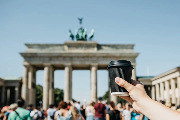 A person holds a disposable cup with coffee or another drink on the background of the Brandenburg Gate in Berlin. - Fotoğraf, Görsel