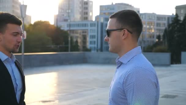 Profile of two young business men standing outdoor and talking. Businessmen meet and speaking outside in the city with blue sky at background. Communication of happy colleagues. Close up Slow motion - Footage, Video