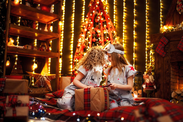 Happy little girls wearing Christmas pajamas open gift box by a fireplace in a cozy dark living room on Christmas eve. - Foto, Imagem