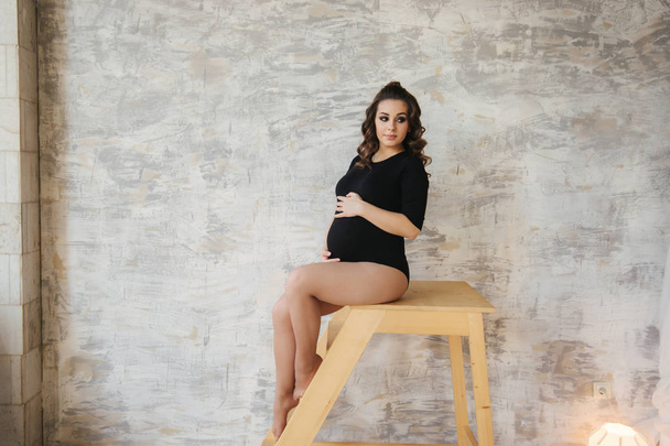Pregnant moman sit on the stairs. Model dressed in black bodysuit. She put her hand on the belly. Happy future mother - Фото, изображение
