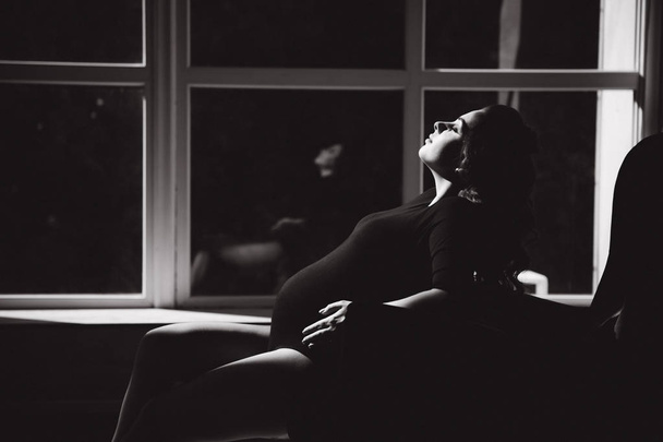 Kaunis raskaana oleva nainen istuu tuolilla edessä iso ikkuna
 - Valokuva, kuva