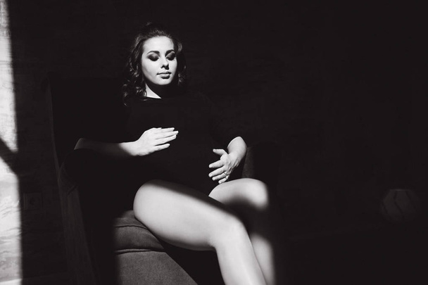 Zwart-wit foto van zwangere vrouw zittend op de stoel. Zon stralen schijnen op een vrouw - Foto, afbeelding