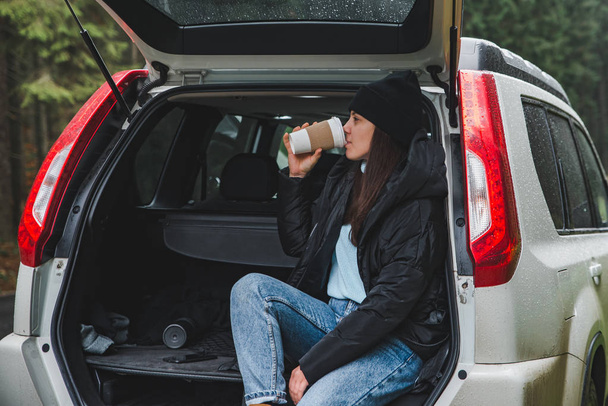young pretty woman sitting in suv car trunk at road side drinking hot coffee at road side in mountain forest road trip concept - Photo, Image