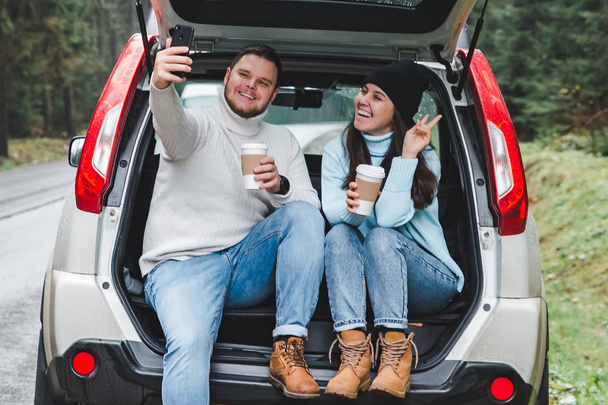 man and woman stop to drink warm up tea and taking selfie in car suv trunk. road trip concept - Zdjęcie, obraz