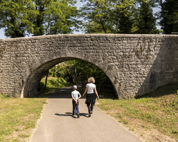 μια γυναίκα με ξανθιά μπούκλες βόλτες με τον γιο της στο δρόμο κάτω από μια πέτρινη γέφυρα - Φωτογραφία, εικόνα