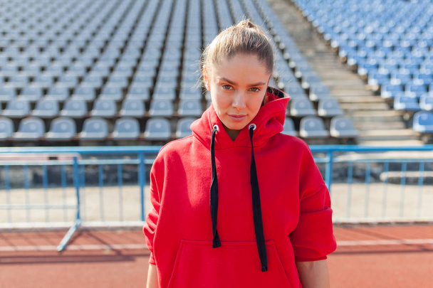 Young sports woman in red sweatshirt resting after jogging training on the running track at summer - Foto, Imagen
