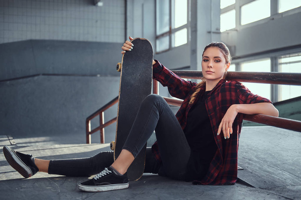 A beautiful young girl wearing a checkered shirt holding a skateboard while sitting next to a grind rail in skatepark indoors. - Photo, Image