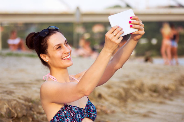 Belle femme souriante en bikini utilise une tablette sur la plage
. - Photo, image