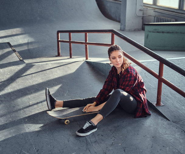 ein schönes junges Mädchen im karierten Hemd hält ein Skateboard in der Hand, während sie im Skatepark neben einer Schleifschiene sitzt. - Foto, Bild