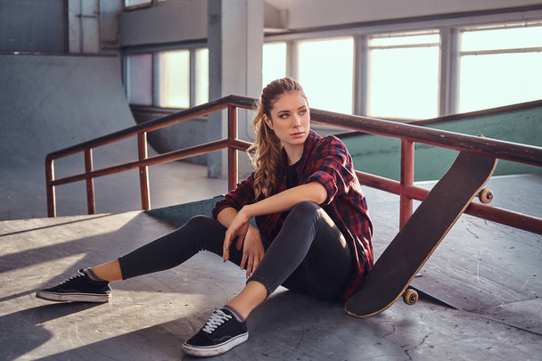 A beautiful young girl wearing a checkered shirt holding a skateboard while sitting next to a grind rail in skatepark indoors. - Foto, imagen