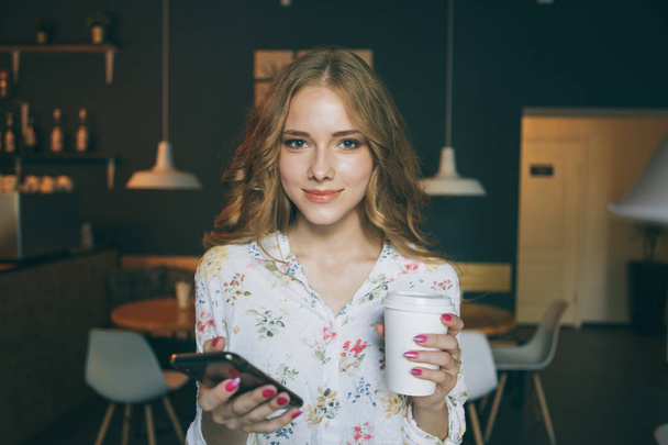 Νεαρή ξανθιά γυναίκα σε μια καφετέρια, ένα κομψό μοντέρνο κορίτσι με καφέ. Φυσάει τα μαλλιά. Νεαρή κοπέλα που κρατά έναν καπουτσίνο και κοιτάζει το κινητό σας τηλέφωνο - Φωτογραφία, εικόνα