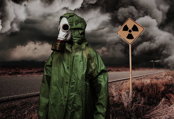 Man in gas mask and cloak for chemical protection on abandoned road with radiation area caution sing under heavy clouds. Nature protection concept - Photo, Image
