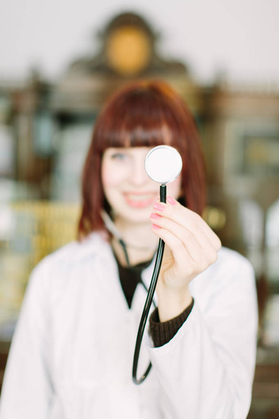 Молодая привлекательная улыбающаяся женщина-фармацевт держит стетоскоп и позирует в зале винтажной аптеки
 - Фото, изображение