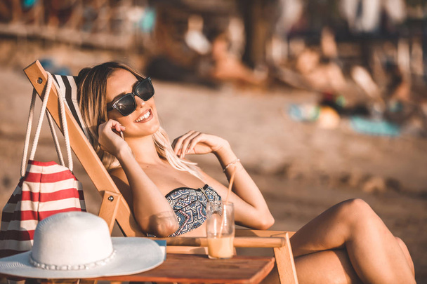 Aantrekkelijke jonge vrouw in een zwembroek en zonnebril zittend op een ligstoel op het strand, praten op een mobiele telefoon en genieten van een mooie zonnige dag - Foto, afbeelding