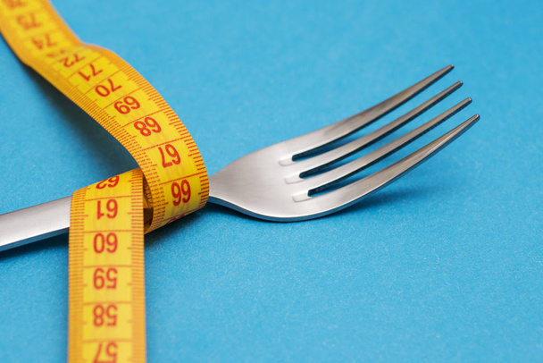 Measuring tape with fork on blue background. Concept of weight loss. Weight management. Healthy lifestyle. Weight loss health issue. Diet concept - 写真・画像