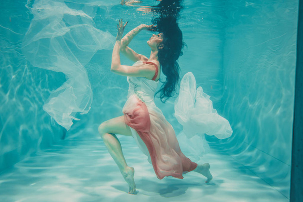 schönes Model-Mädchen schwimmt unter Wasser in femininem langen weißen und rosa Kleid - Foto, Bild