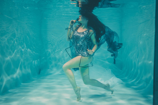 piękny model dziewczyna pływa pod wodą w t-shirt szary cekinami i przejrzyste modny płaszcz PCV - Zdjęcie, obraz