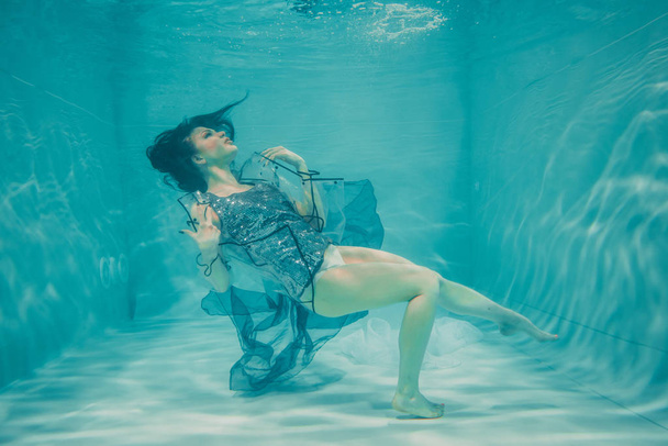το όμορφο μοντέλο κορίτσι κολυμπάει κάτω από το νερό σε ένα μπλουζάκι σε γκρι παγιέτες και ένα διαφανές αδιάβροχο Pvc μόδας - Φωτογραφία, εικόνα