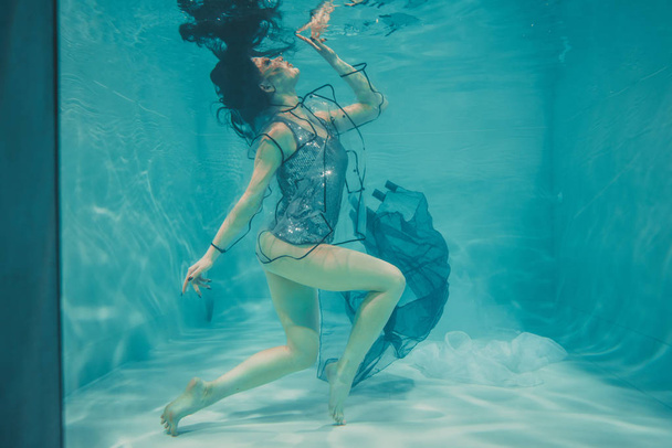 beautiful model girl swims under water in a t-shirt in gray sequins and a transparent fashionable PVC raincoat - Photo, Image