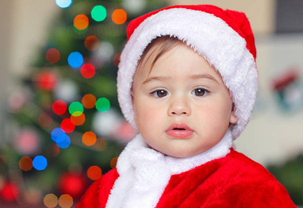 Portrait of a beautiful little Santa Claus at home, sweet baby boy dressed in red festive costume with pleasure playing near Christmas tree - Valokuva, kuva
