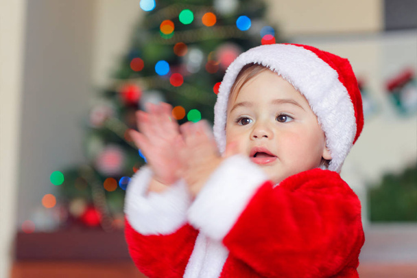 Retrato de um menino bonito feliz se divertindo em casa na véspera de Natal, criança adorável vestindo traje de Papai Noel batendo palmas perto da árvore de Natal - Foto, Imagem
