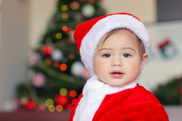 Portrait of a beautiful little Santa Claus at home, sweet baby boy dressed in red festive costume with pleasure playing near Christmas tree - Фото, изображение
