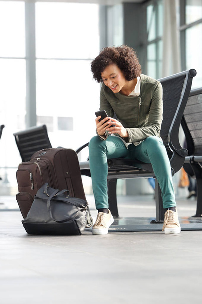 Portret african american kobieta siedzi na ławce na stacji z bagażem i telefonu komórkowego - Zdjęcie, obraz