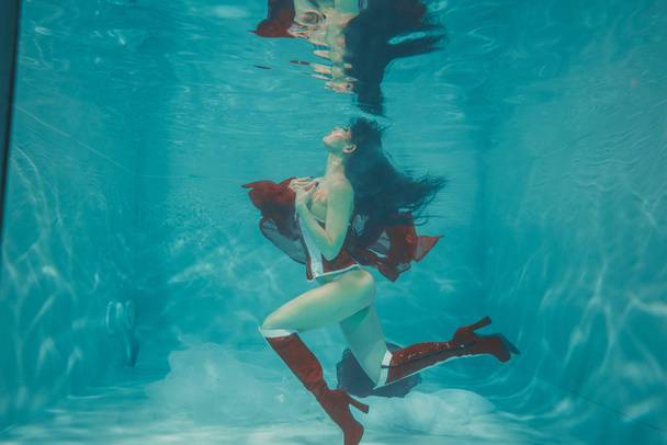 mooi sexy meisje onderwater zwemmen in sportieve stijl rode en witte fetish korset en over knie dij hoge laarzen met hakken - Foto, afbeelding