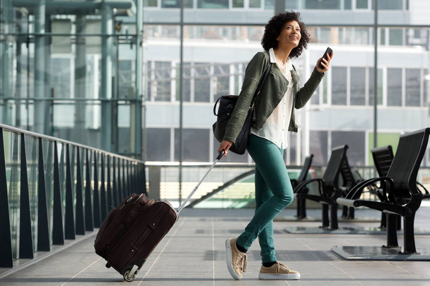 Retrato lateral de cuerpo completo de mujer viajera caminando en la estación con maleta y teléfono celular
  - Foto, imagen